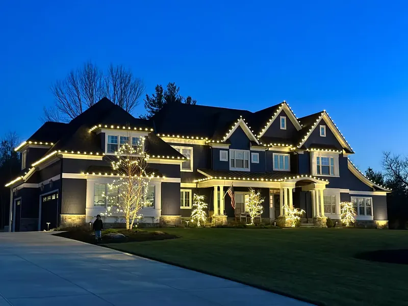 Two-story home at dusk with warm white Christmas lights outlining its rooflines and entrance
